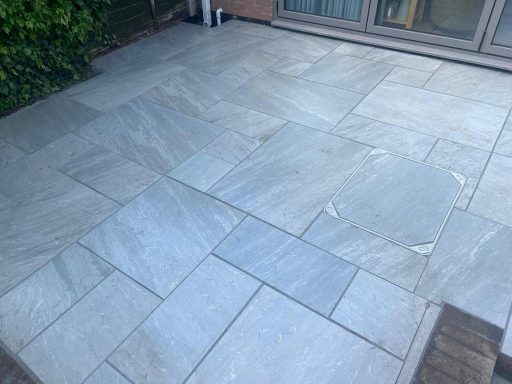 A paved patio with large grey stone slabs, partially surrounded by greenery.