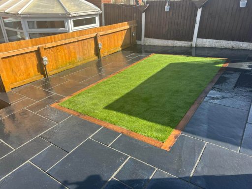 Garden area with artificial grass bordered by paved stone and wooden fencing.