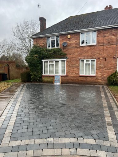 Two-storey brick house with a driveway and greenery, under a cloudy sky.