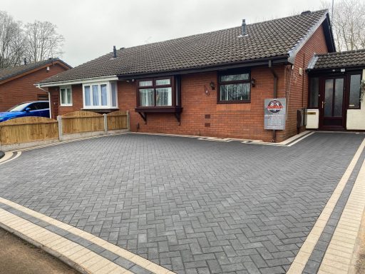 Bungalow with grey block paving and surrounding garden, featuring a driveway and windows.