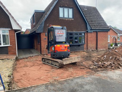 Small digger parked on a driveway beside a house with construction debris around.
