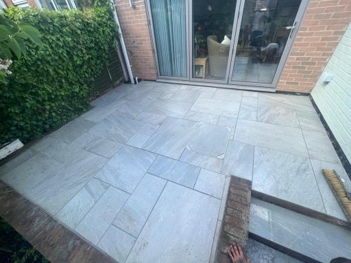 Paved patio area with stone slabs, bordered by greenery and sliding glass doors.