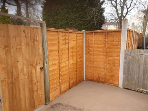 Wooden garden fence corner with a gate, surrounded by greenery.