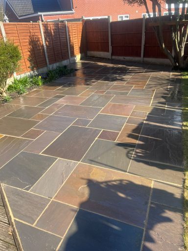 A sunlit garden patio with multi-coloured stone paving and a surrounding wooden fence.