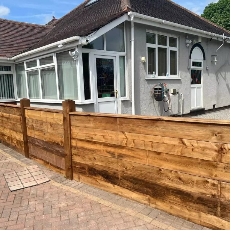 A wooden fence surrounds a light grey bungalow on a paved driveway.
