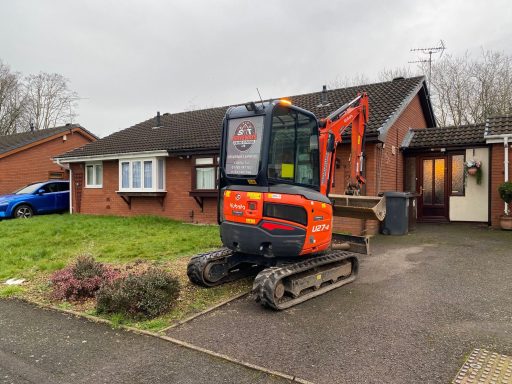 Excavator parked outside a bungalow with a blue car nearby.