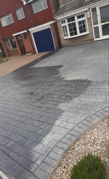 Unevenly coloured patterned driveway with a nearby garden and house facade.