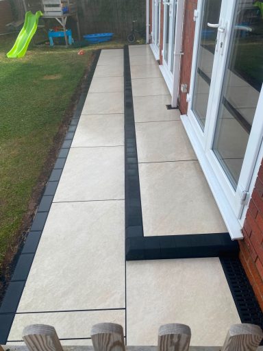 Paved pathway beside a house, featuring light tiles and dark border, leading to a garden.