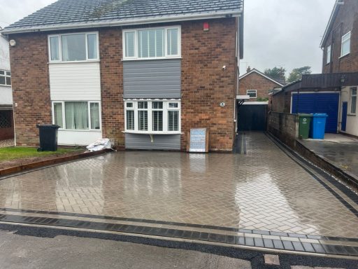 A driveway in front of a two-storey house, featuring wet paving and several bins nearby.