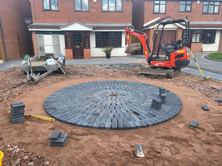 Construction of a circular stone patio with a digger and materials nearby.