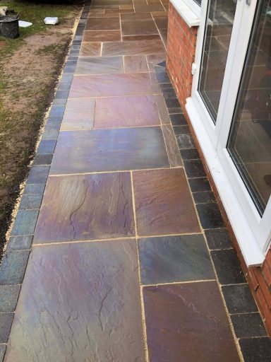 Pathway made of multi-coloured slate tiles beside a brick wall and glass doors.