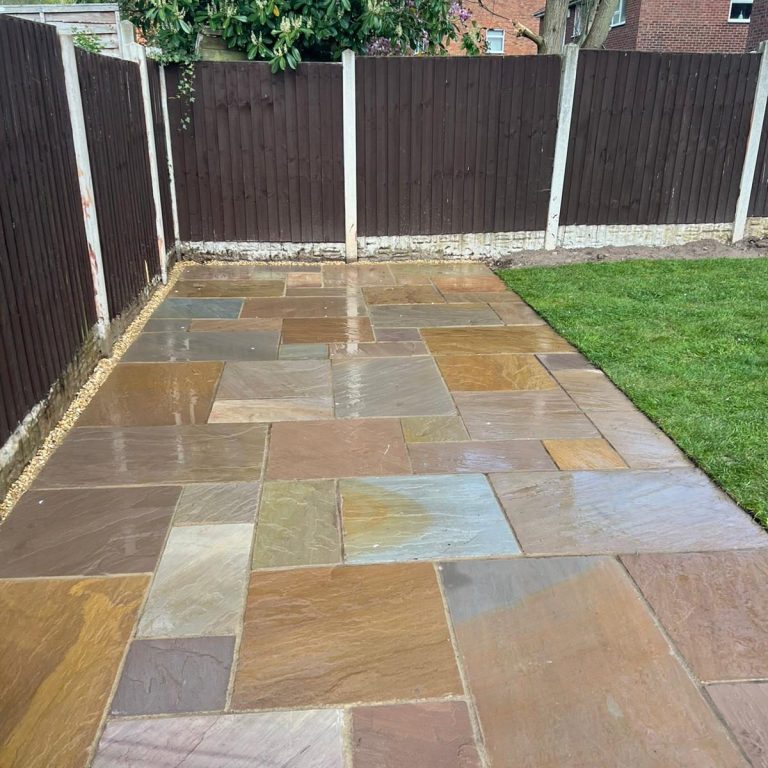 A stone patio with a mix of colours, bordered by wooden fencing and a grassy area.