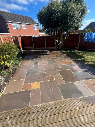A paved garden area with multi-coloured stones and a wooden edge, surrounded by fences and plants.